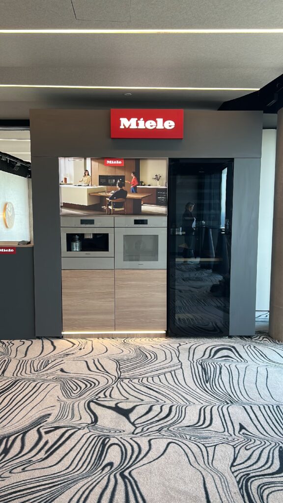W nowoczesnej ekspozycji marki Miele prezentowane są wbudowane urządzenia kuchenne w szafkach w kolorze drewnianym i szarym, duży ekran przedstawiający kuchnię, czerwone logo Miele umieszczone powyżej oraz wzorzysta wykładzina dywanowa. Szklane ściany odbijają postać stojącą w tle. projekt i nadzór Tatjana Kobuszewska