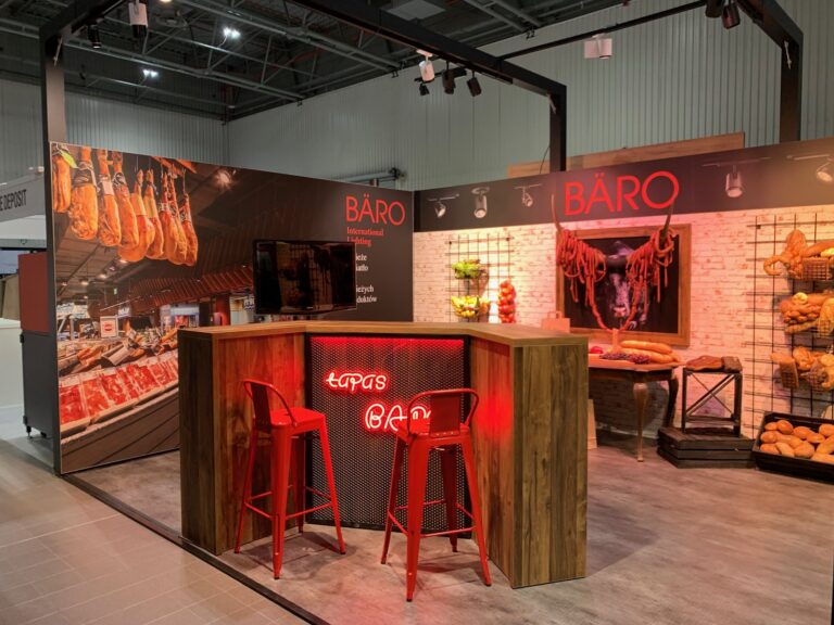 Stoisko oświetleniowe urządzone jak tapas bar: drewniana lada, czerwone hokery, wiszące wędliny i ściana z cegły
