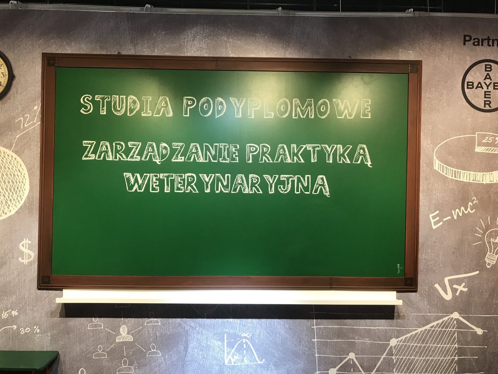 Na zielonej tablicy z drewnianą ramą widnieje napis wykonany białą kredą: STUDIA PODYPLOMOWE ZARZĄDZANIE PRAKTYKĄ WETERYNARYJNĄ. Tablica jest przymocowana do ciemnoszarej ściany, ozdobionej delikatnymi rysunkami symboli matematycznych i naukowych wykonanymi białą kredą. projekt i nadzór Tatjana Kobuszewska