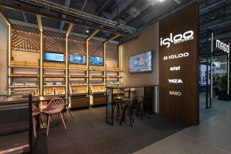 Nowoczesna ekspozycja sklepowa z drewnianymi półkami, ekranami LED oraz efektownym kasetonem z folii 3D z logo „igloo technika”. Krzesła z wikliny, wysokie stołki i stoły zachęcają gości do zanurzenia się w jasnej atmosferze targów Retail Show 2024. projekt i nadzór Tatjana Kobuszewska