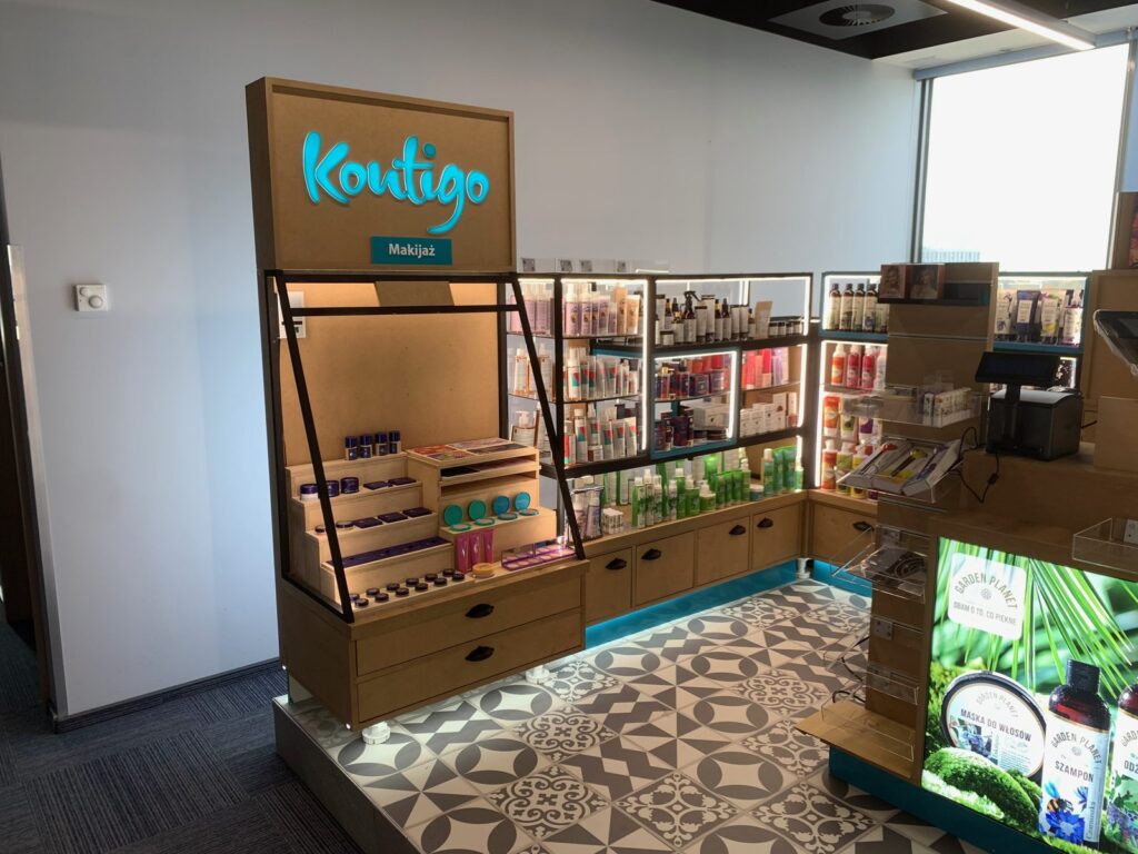 Wieża makijażowa wyspy Kontigo: podświetlane logo z tabliczką „Makijaż”, drewniane ekspozytory schodkowe z cieniami i pomadkami, szuflady pod ladą, czarna rama stalowa. projekt i nadzór Tatjana Kobuszewska trksystem.com