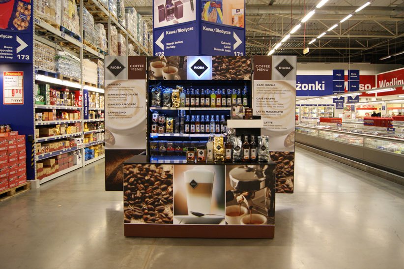 Wyspa Rioba od frontu: podświetlany kaseton z logo, półki z taśmami LED zatowarowane kawą i syropami, po bokach panele z wydrukowanym menu kawowym, cokół z fotografią ziaren.