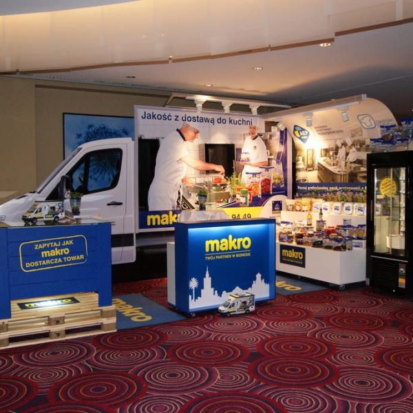 Stoisko targowe z brandingiem „makro”, zaprojektowane przez projekt i nadzór trksystem.com, obejmuje furgonetkę, szefa kuchni przygotowującego jedzenie, eksponowane produkty spożywcze, lodówkę, materiały promocyjne i duże polskie tło ustawione na czerwonym dywanie. projekt i nadzór Tatjana Kobuszewska trksystem.com