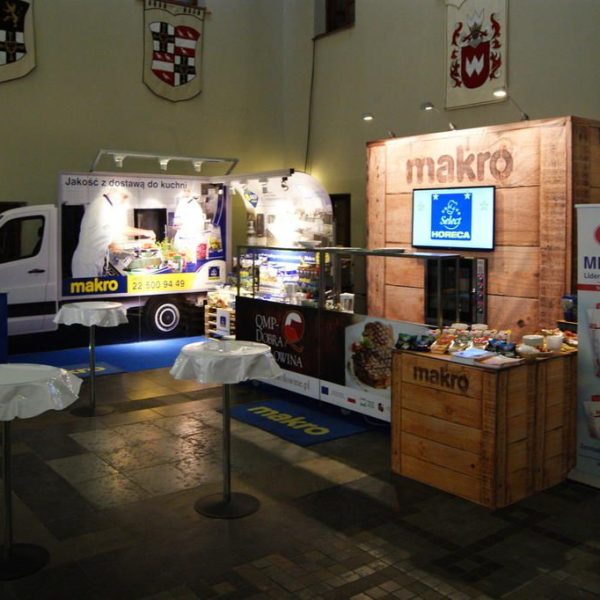 Stoisko targowe prezentuje markę Makro, tło w postaci samochodu dostawczego, konstrukcje w stylu skrzyń i banery Mlekovita w hali z kafelkową podłogą i herbem, na której znajduje się projekt i nadzór trksystem.com autorstwa Tatjany Kobuszewskiej. projekt i nadzór Tatjana Kobuszewska trksystem.com