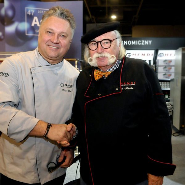 Kurt Scheller i Robert Sowa Dwóch mężczyzn w strojach szefów kuchni ściska sobie dłonie i uśmiecha się do siebie podczas Forum Szefów Kuchni i restauratorów w Expo XXI, w tle widać sprzęt kuchenny i baner Hendi. projekt i nadzór Tatjana Kobuszewska trksystem.com