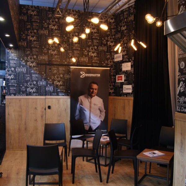 Przytulny kącik kawiarniany z czarnymi krzesłami wokół małych stolików na podłodze z jasnego drewna. To część stoiska targowego Hendi na Targach SweetProjekt Tatjana Kobuszewska, projekt i nadzór trksystem.com. Nowoczesne lampy wiszące i tablice kredowe tworzą ciepłą, zachęcającą atmosferę. projekt i nadzór Tatjana Kobuszewska trksystem.com