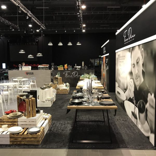 Nowoczesny showroom ze starannie ustawionymi stołami, na których eksponowana jest elegancka zastawa stołowa, szklanki i sztućce. Białe krzesła i skrzynki dodają uroku wystrojowi. Na otaczających czarnych ścianach wiszą duże czarno-białe zdjęcia szefa kuchni. projekt i nadzór Tatjana Kobuszewska trksystem.com