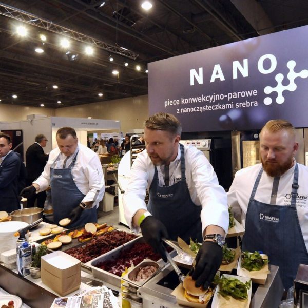 Trzech kucharzy w białych uniformach i czarnych rękawiczkach przygotowuje jedzenie na ruchliwym wydarzeniu lub targach. Za nimi na szyldzie widnieje napis NANO dużymi literami, z mniejszym tekstem po polsku. Ludzie patrzą w tle. projekt i nadzór Tatjana Kobuszewska trksystem.com