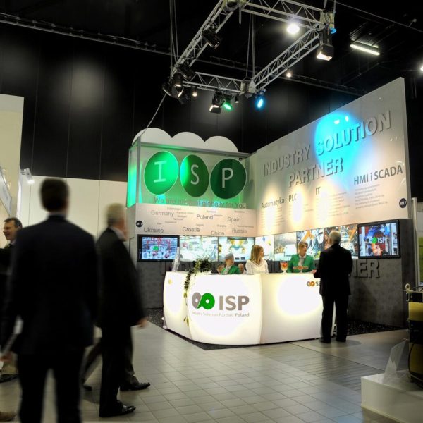 Jasno oświetlone stoisko targowe ISP, Industry Solution Partner, z personelem za ladą, wieloma ekranami i dużymi zielonymi okrągłymi znakami. Kilka osób przechodzi lub wchodzi w interakcję w ruchliwej hali wystawowej. projekt i nadzór Tatjana Kobuszewska trksystem.com