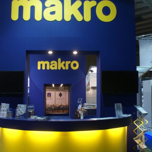 Stoisko targowe z niebieskimi ścianami i dużymi żółtymi logotypami makro, z zakrzywionym biurkiem recepcji ze świecącym żółtym frontem, broszurami i dwoma płaskimi ekranami zamontowanymi na ścianie. projekt i nadzór Tatjana Kobuszewska trksystem.com