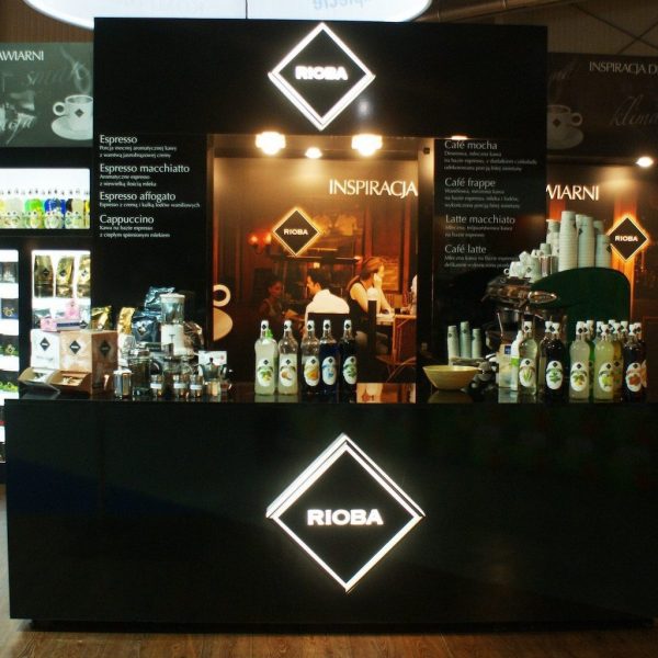 Eleganckie czarne stoisko z kawą z logo „RIOBA”, ekspresy do kawy, butelki i kubki na ladzie. Podświetlane menu zawierają listę napojów espresso; po lewej stronie widoczna jest lodówka, a za nią tło w stylu kawiarnianym. projekt i nadzór Tatjana Kobuszewska trksystem.com