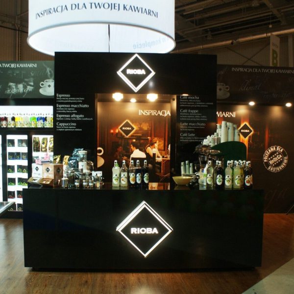 Nowoczesne stoisko z kawą z logo „RIOBA”, podświetlanymi szyldami i różnymi produktami kawowymi; barista pracuje za ladą na targach EuroGastro. projekt i nadzór Tatjana Kobuszewska trksystem.com