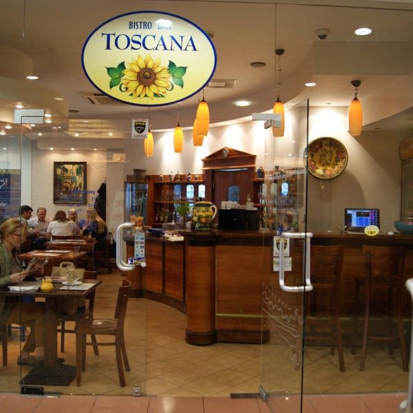 Przeszklone bistro o nazwie "Toscana" prezentuje projekt restauracji z ciepłym oświetleniem, drewnianymi meblami i klientami spożywającymi posiłki w środku. Wejście zdobią wiszące lampy, logo słonecznika nad drzwiami i odręczne menu na szybie. projekt i nadzór Tatjana Kobuszewska trksystem.com