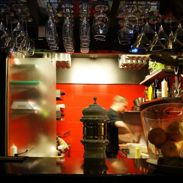 Szef kuchni pracuje w nowoczesnej kuchni baru tapas Carmona z odważnymi czerwonymi kafelkami, wiszącymi szklanymi naczyniami, półkami zaopatrzonymi w składniki i ladą z owocami i chlebem na pierwszym planie. projekt i nadzór Tatjana Kobuszewska trksystem.com