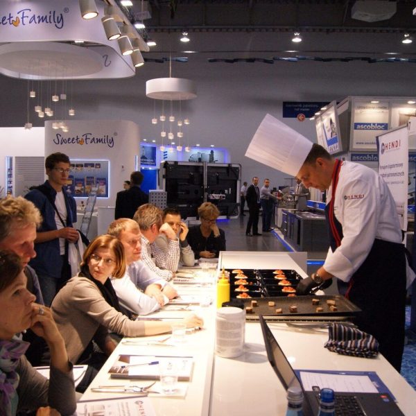 Szef kuchni w wysokiej białej czapce przygotowuje desery na blacie, podczas gdy grupa osób przy stole uważnie obserwuje pokaz gotowania Hendi na targach Polagra. projekt i nadzór Tatjana Kobuszewska trksystem.com