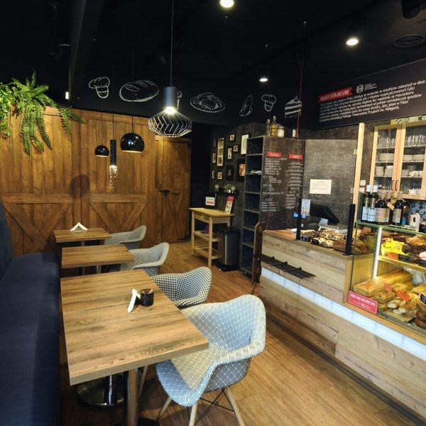 Przytulna "piekarnia przystanek" kawiarnia z drewnianymi stołami, wyściełaną ławą, wzorzystymi krzesłami, ścianą z oprawionymi zdjęciami, wiszącymi roślinami oraz witryną wypełnioną ciastami i pieczywem. Ciepłe oświetlenie tworzy zachęcającą atmosferę. projekt i nadzór Tatjana Kobuszewska trksystem.com