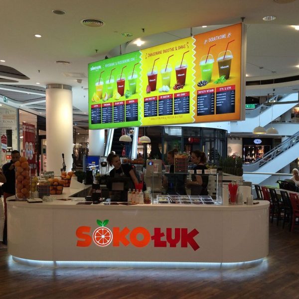 tętniąca życiem wyspa gastronomiczna Redberry-Sokołyk w centrum handlowym, z personelem za ladą, wystawami owoców i kolorowymi tablicami z menu powyżej. W tle widoczni są klienci i część wypoczynkowa. projekt i nadzór Tatjana Kobuszewska trksystem.com