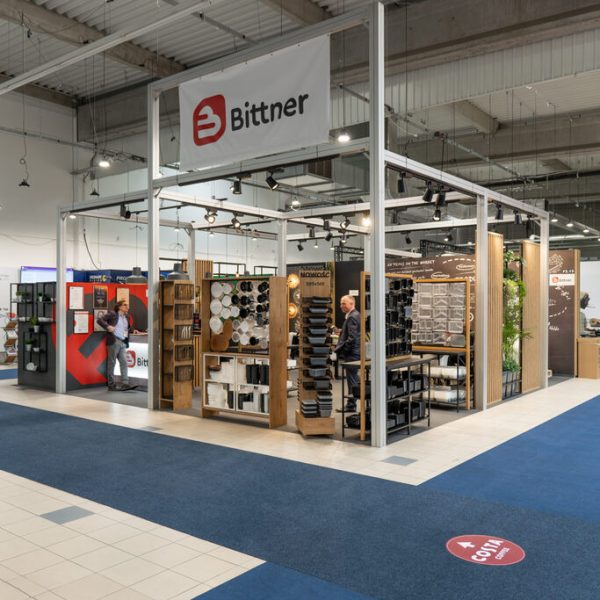 Stoisko targowe dla firmy Bittner, wyposażone w regały z różnymi produktami sprzętowymi i ekspozytory, ustawione w dużej hali wystawienniczej z niebieską wykładziną i industrialnym sufitem. Projekt i nadzór wykonawczy zapewniły precyzyjne wykonanie i Foto: Tatjana Kobuszewska.
